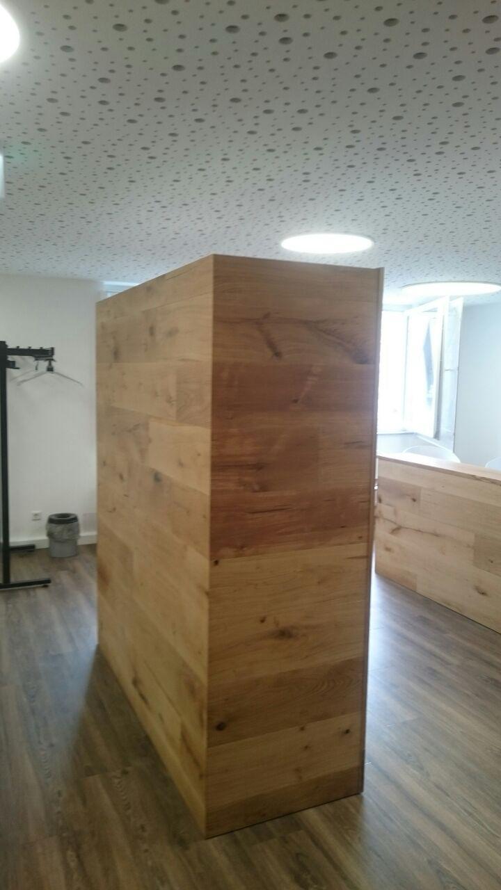 Ein großer Holzquader steht auf dunklem Boden in einem hellen Raum mit Lochdecke