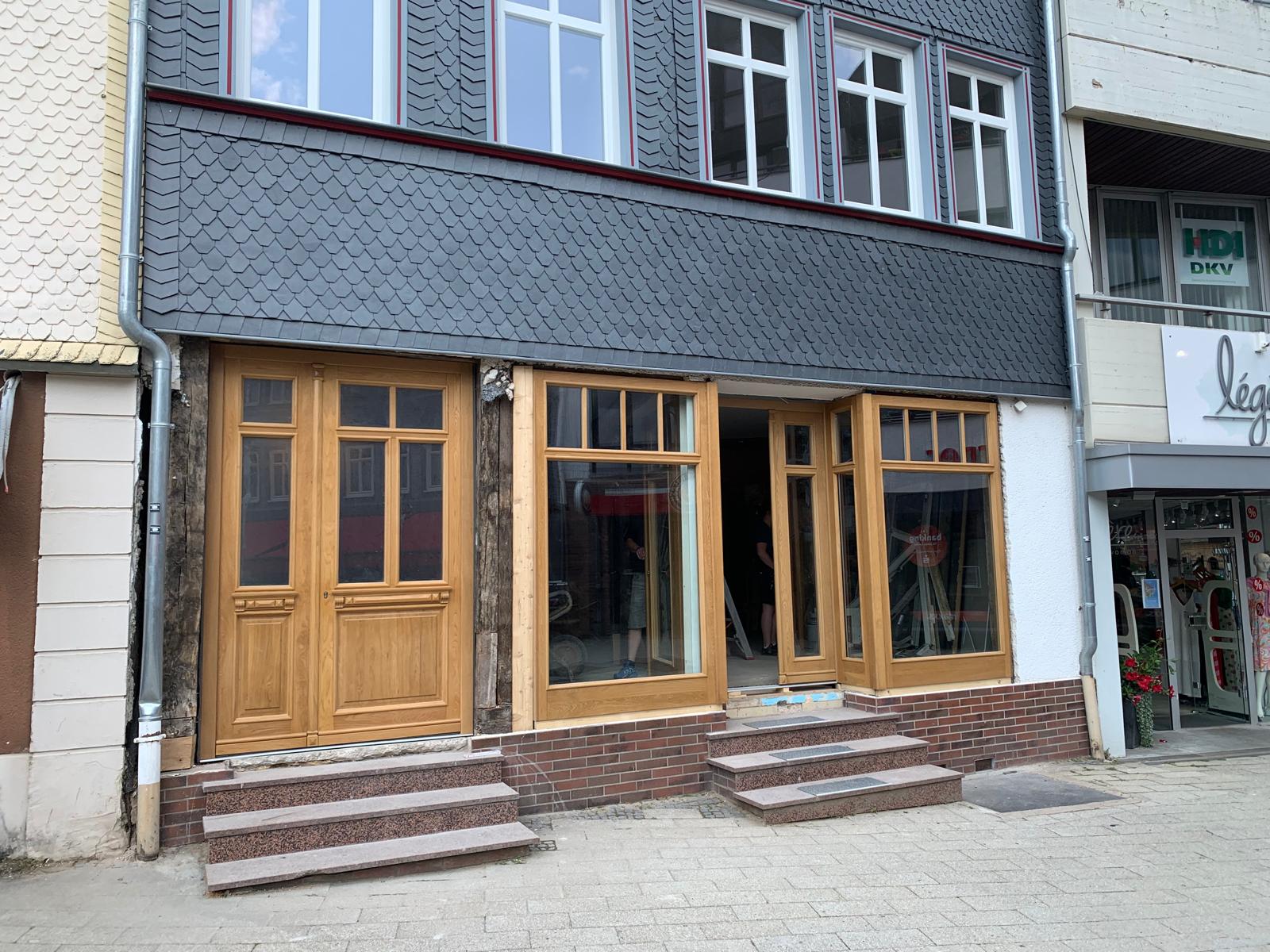 Zwei offene Holztüren mit großen Glasflächen an einem grauen Gebäude mit Treppenstufen